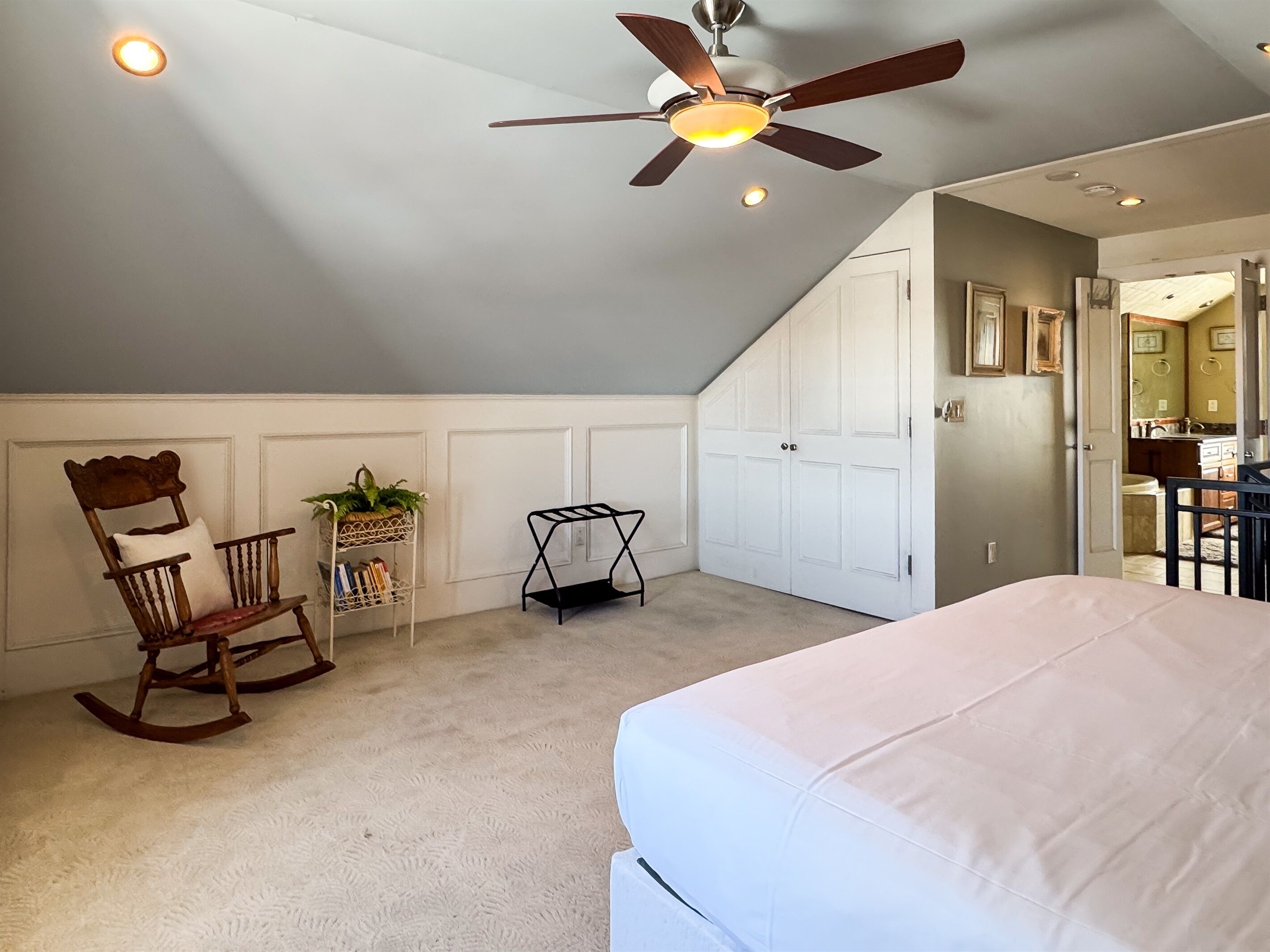 Un dormitorio con cama, sillón mecedor, planta y ventilador de techo.