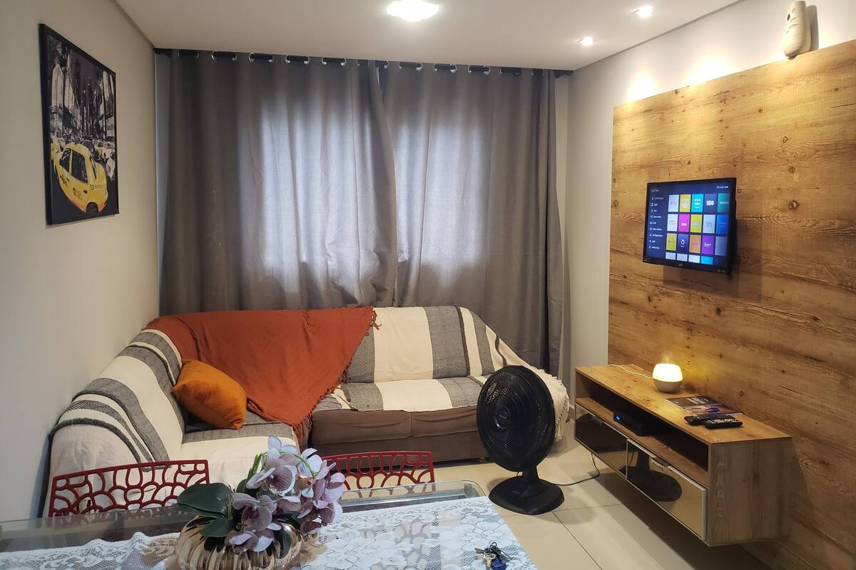 A modern living room with a sofa, a TV mounted on a wooden wall, a fan, and a table with a vase of flowers.
