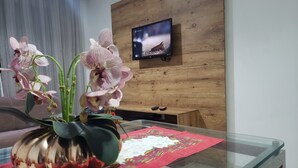 Een woonkamer met een houten wand, een tv die eraan bevestigd is, een glazen salontafel met een rode tafelonderzetter en een orchidee in een pot.