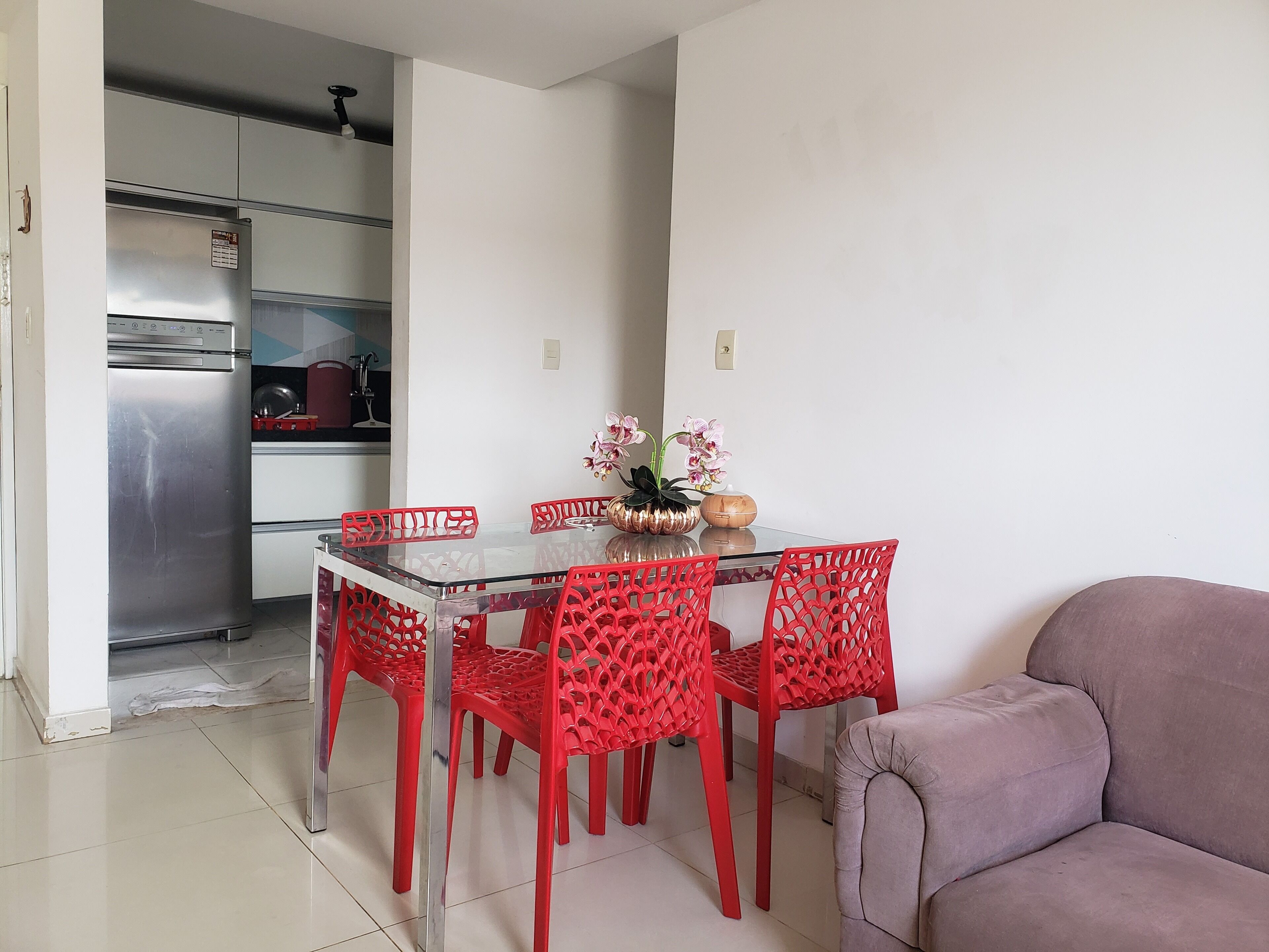 A dining area with a glass table and red chairs, a kitchen in the background, and a sofa to the right.