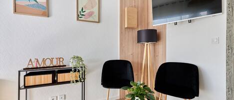 A modern living room with a wooden floor, a black TV mounted on the wall, two black chairs, a round wooden table with a plant, and a black floor lamp.