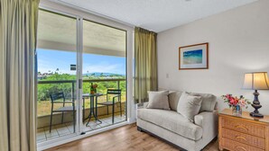 A room with a sliding glass door to a balcony, a sofa, a small table with chairs, a wicker side table with a lamp, and a framed picture on the wall.