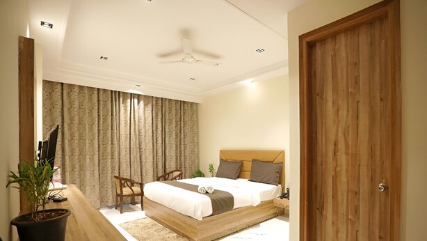 A bedroom with a bed, wooden headboard, curtains, a desk with a plant, a chair, and a ceiling fan. - Hotel GoodLuck House Near Delhi Airport (New Delhi)