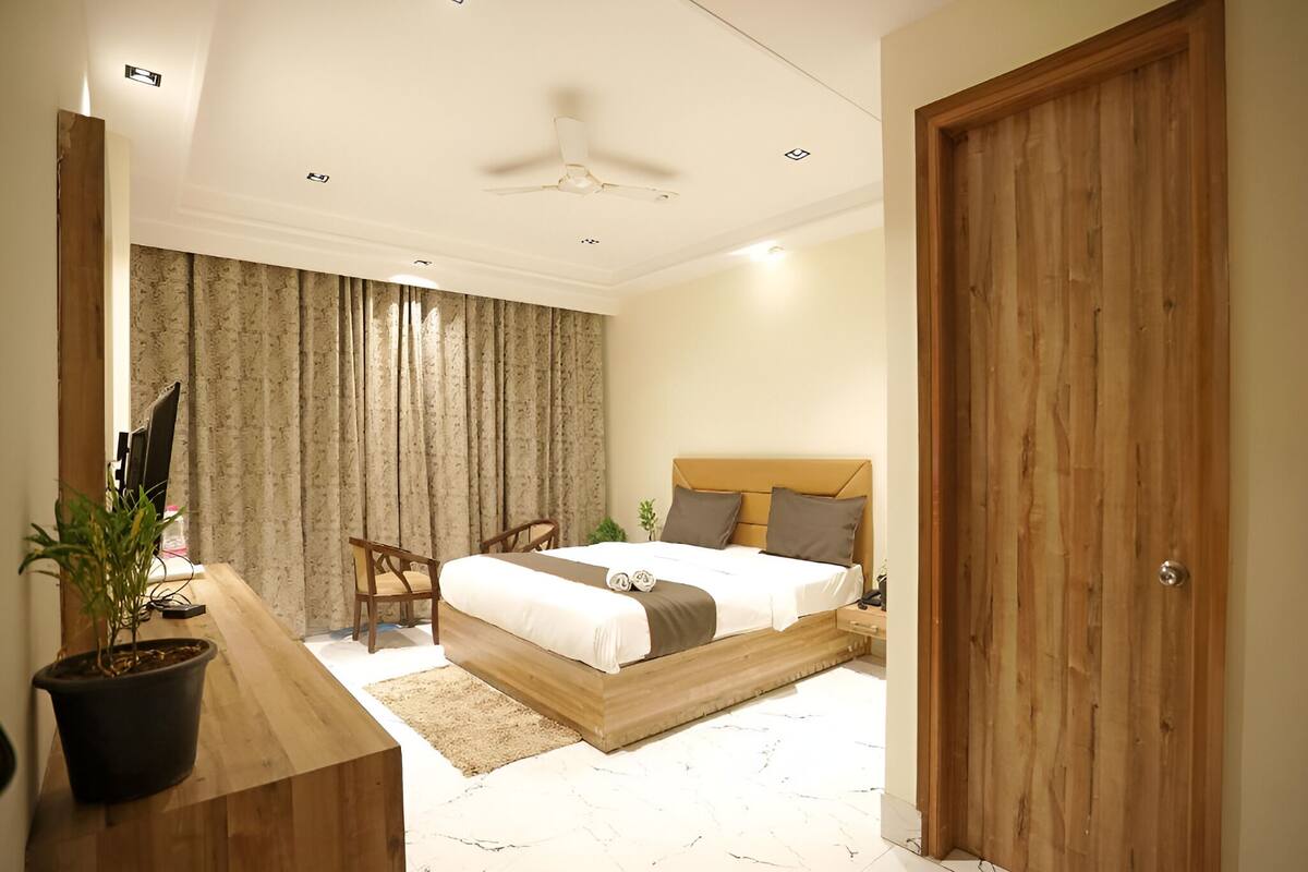 Une chambre à coucher avec un lit, une tête de lit en bois, des rideaux, un bureau avec une plante, une chaise et un ventilateur de plafond.