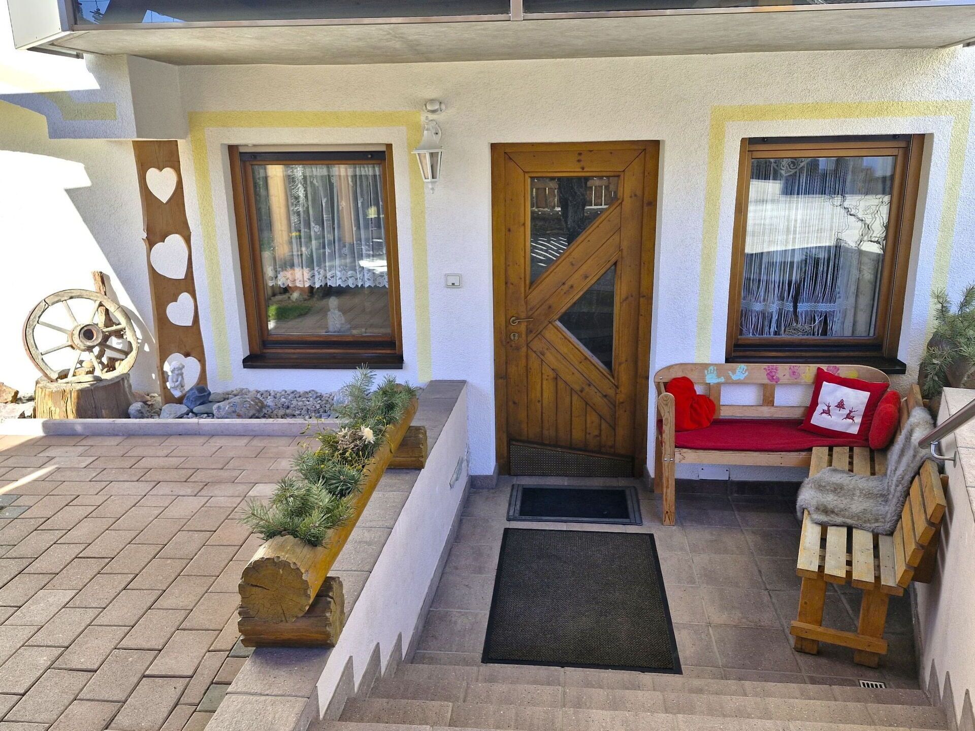 Une entrée avec une porte en bois, un banc doté d’un coussin rouge et un bac à fleurs orné de plantes vertes.