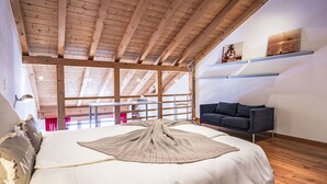 Ein geräumiges Zimmer mit hohen Decken, Holzbalken, einem großen Bett mit gemustertem Überwurf, einer dunklen Couch und Blick ins Freie durch große Fenster.