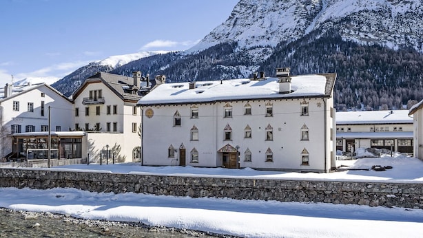 Ein mit Schnee bedecktes Gebäude mit einer Steinwand im Vordergrund, vor einer Bergkulisse.