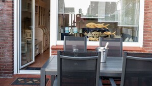 Un área de estar al aire libre con una mesa y sillas, un ventanal de vidrio que da a un espacio interior, y peces decorativos en la ventana.