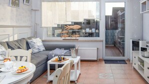 Una sala de estar moderna con un sofá gris, una mesa de comedor con platos y vasos, una pequeña mesa blanca con una bandeja, y vista a una cocina a través de un ventanal grande.