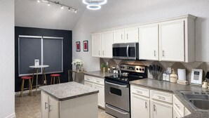 A modern kitchen with white cabinets, a stainless steel stove, and a microwave. There are two stools and a small table with a vase of flowers. - Luxury Couples Hideaway | King Bed • Jacuzzi Spa • Private Retreat (Killeen)