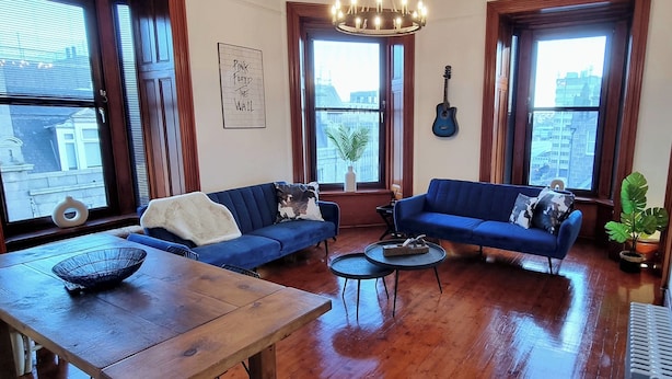 Un salón amplio con pisos de madera, dos sofás azules, una mesa de comedor con un bol y ventanas grandes.