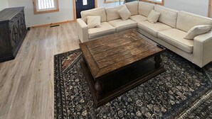 Un salon spacieux avec un plafond haut, des poutres apparentes, un grand canapé en forme de L, une table basse et un tapis à motifs.