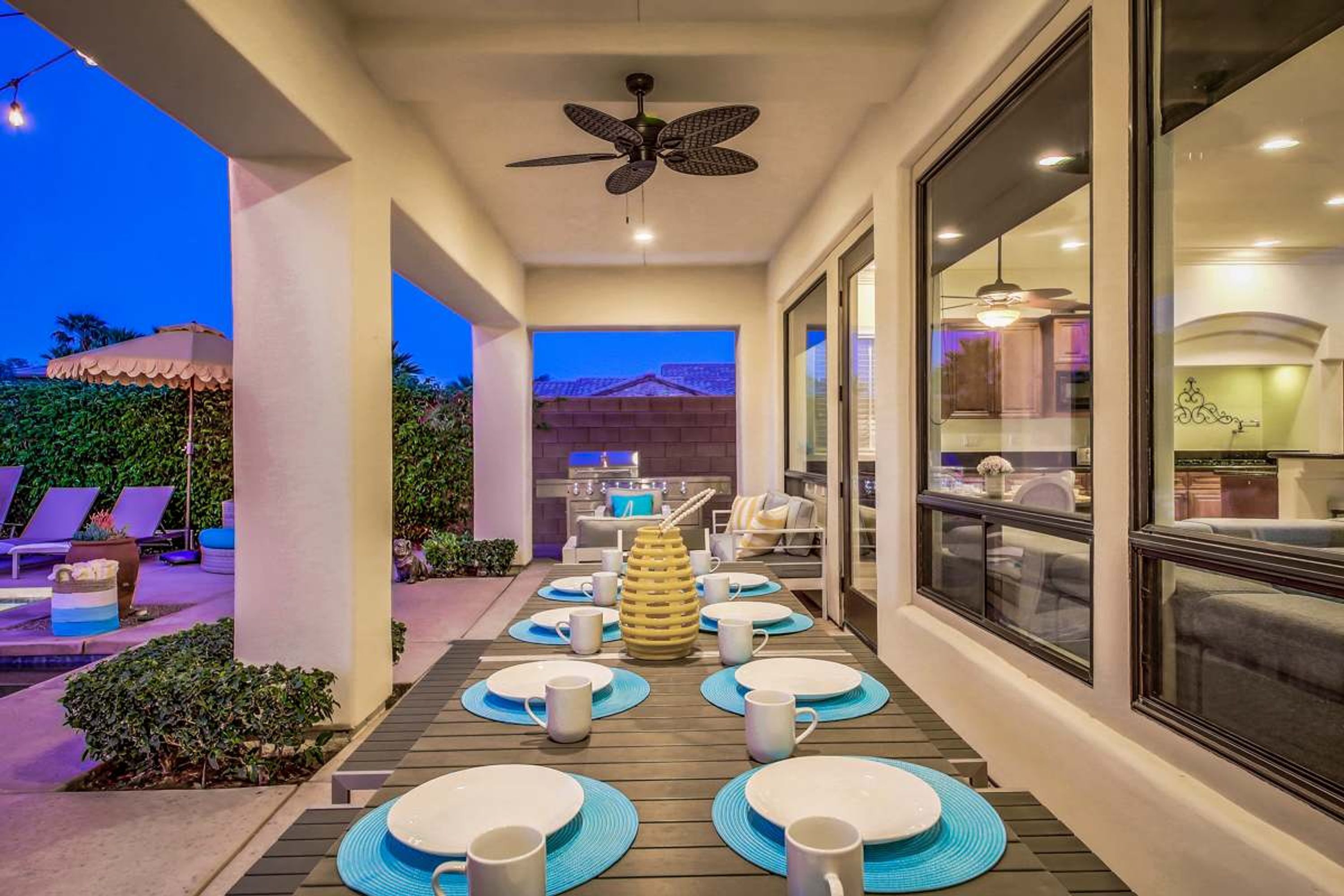 Un espace de restauration en plein air avec une longue table dressée pour accueillir plusieurs personnes, un ventilateur de plafond et une vue sur la cuisine et les montagnes en arrière-plan.