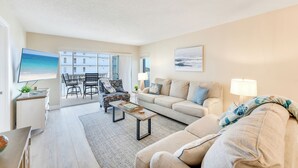 A living room with a sofa, coffee table, and TV. There is a balcony with a view of buildings and a beach.