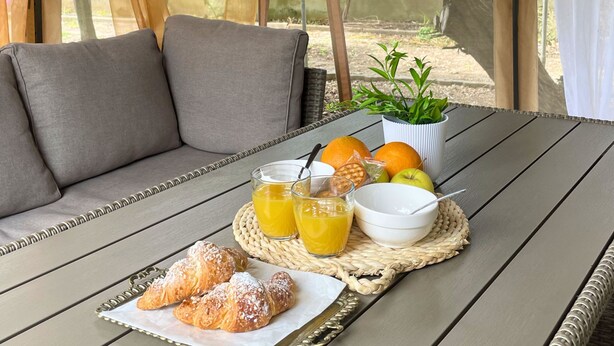 A covered outdoor seating area with a table set for breakfast, including croissants, orange juice, and a bowl of fruit.