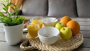 A breakfast setup with a woven placemat, a potted plant, a bowl, two glasses of orange juice, and fruits on a wooden table.