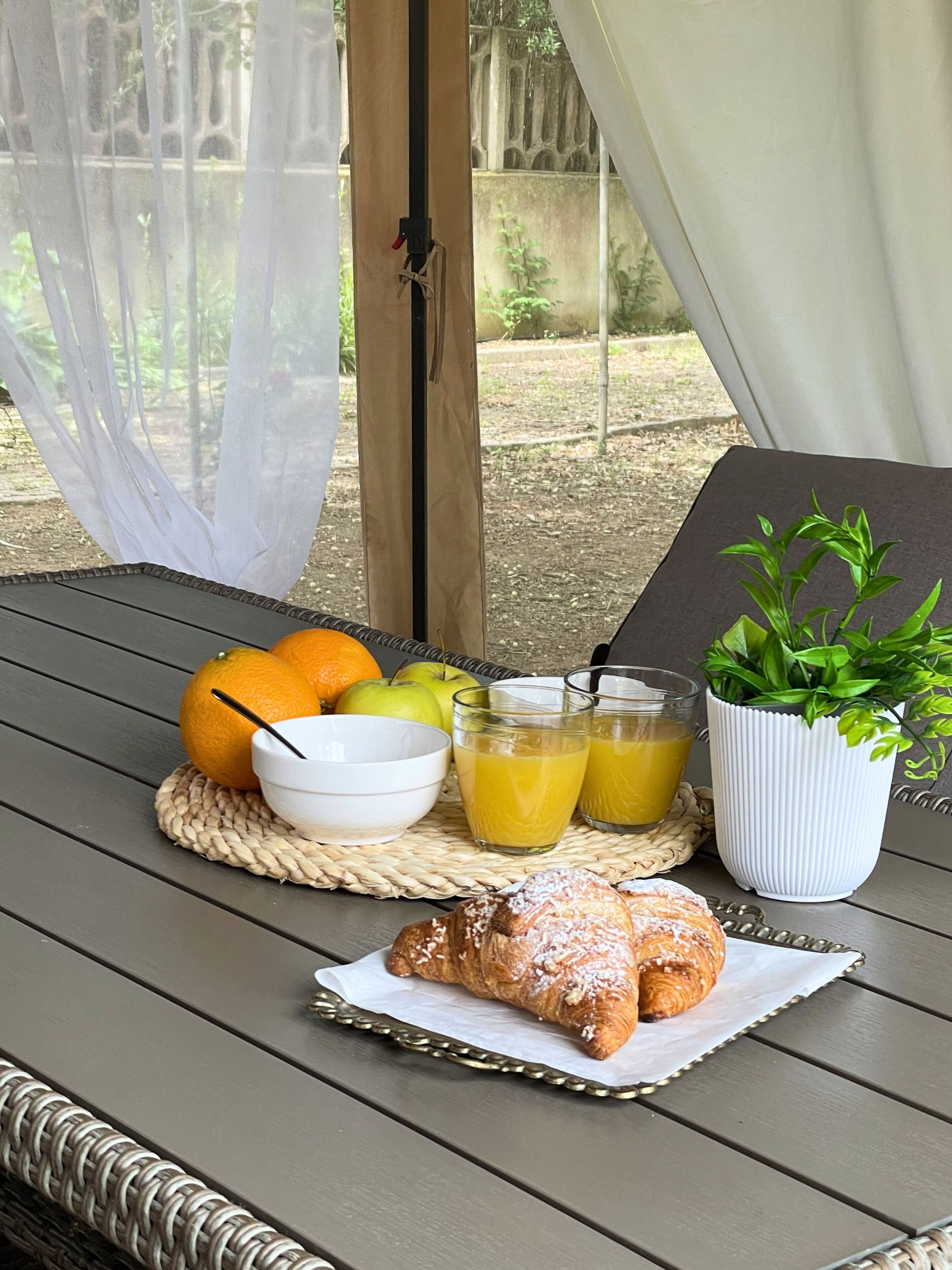 A table with breakfast items including fruits, juice, and pastries, set on a wicker table with a view of a garden.