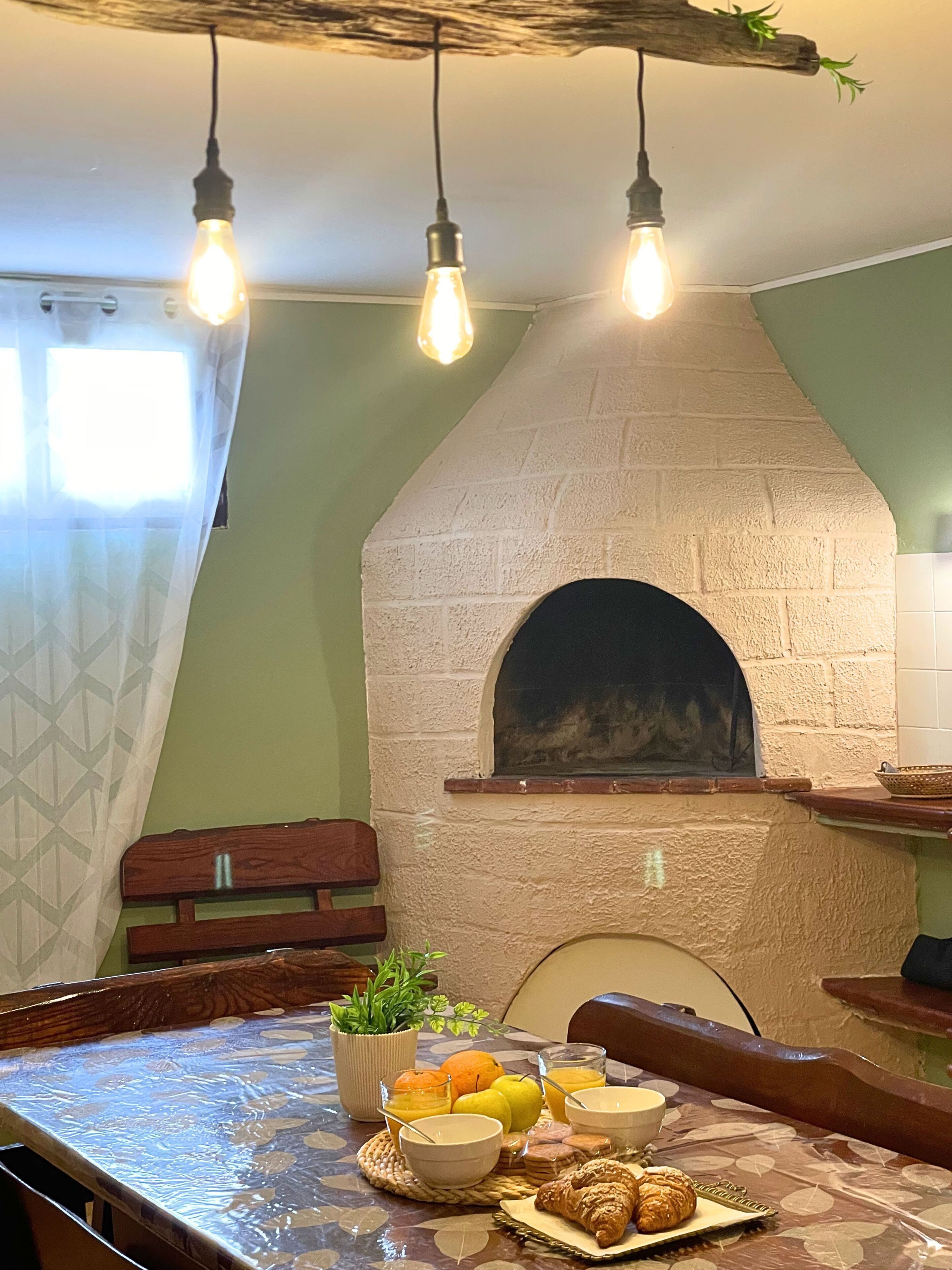 A kitchen with a brick oven, hanging light bulbs, a table with breakfast items, and a window with a curtain.