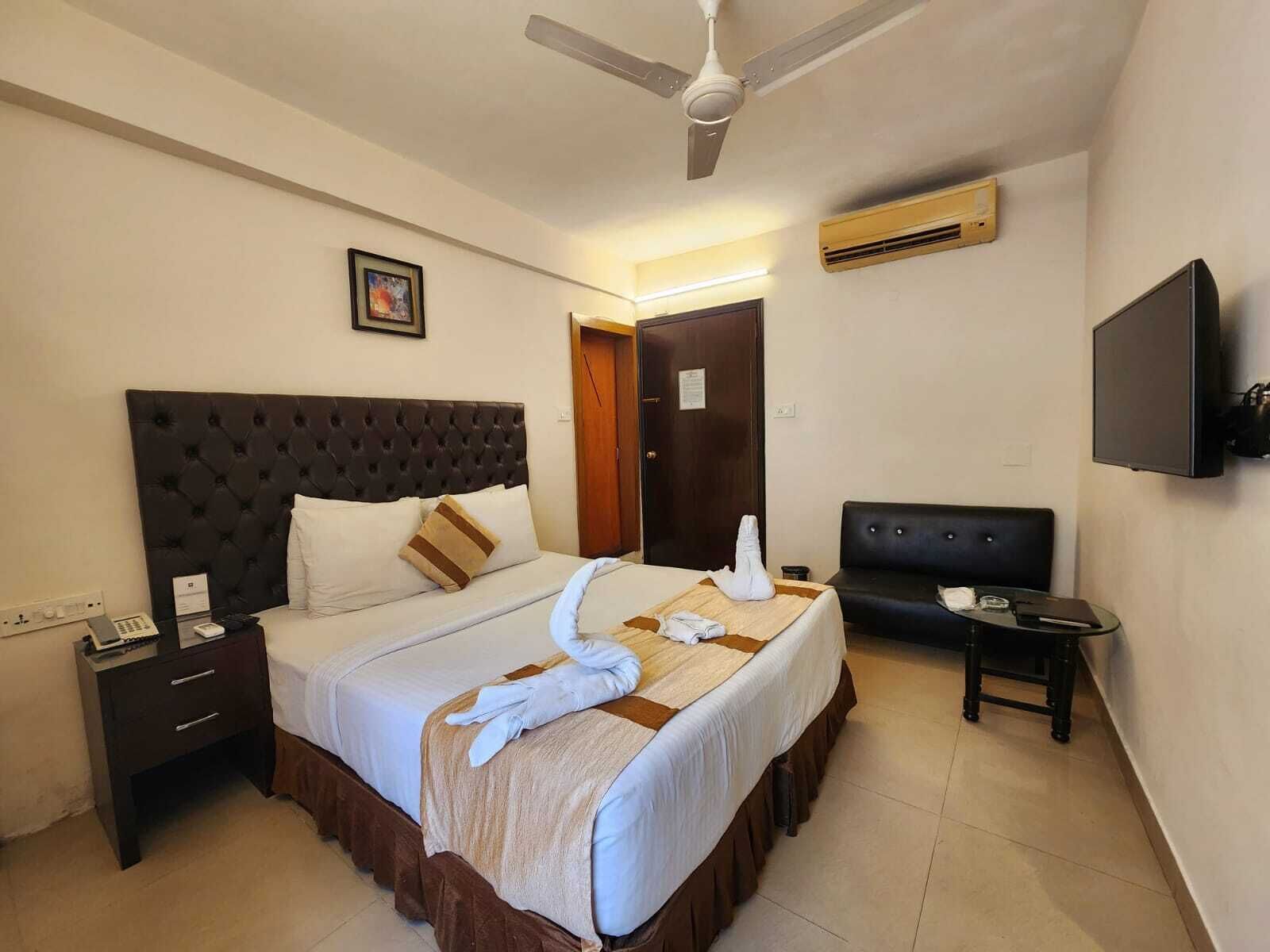 A hotel room with a large bed, a bedside table, a desk, a chair, a TV, and an air conditioning unit.