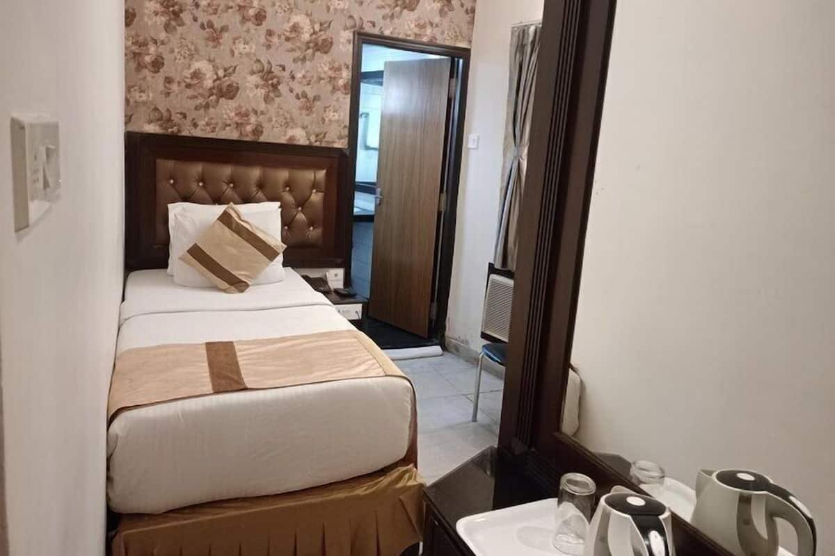 A hotel room with a bed, a wooden headboard, a floral wallpaper, a door, and a small table with a kettle and cups.
