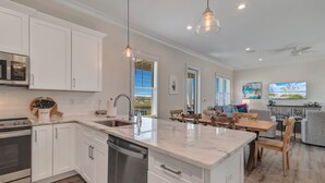 A modern kitchen with white cabinetry, a central island, and a dining area with wooden furniture. - Azure Horizon Beachfront Charm in Gulf Shores (Gulf Shores)