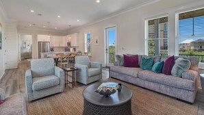 A spacious living room with a sofa, chairs, and a coffee table. A kitchen is visible in the background with a dining area. - Azure Horizon Beachfront Charm in Gulf Shores (Gulf Shores)