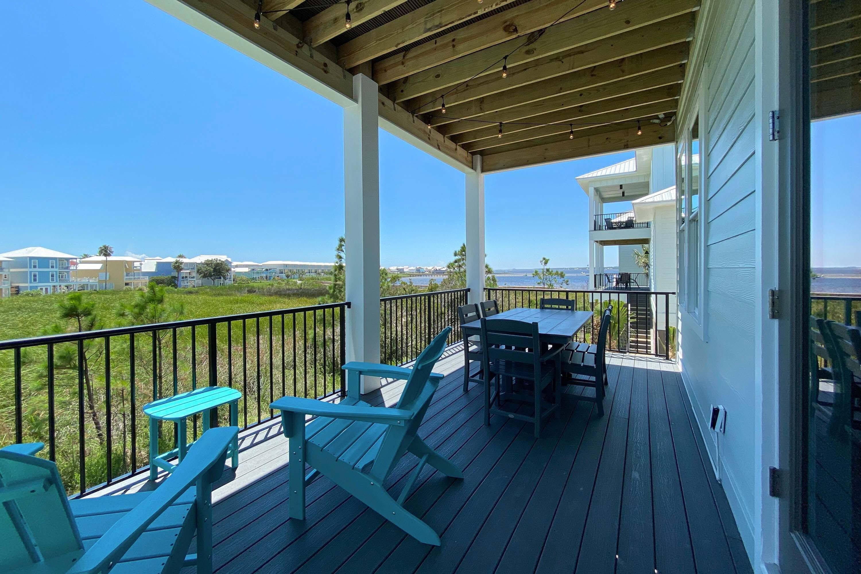 A wooden deck with outdoor furniture, a table, and chairs, overlooking a grassy area and buildings in the distance.