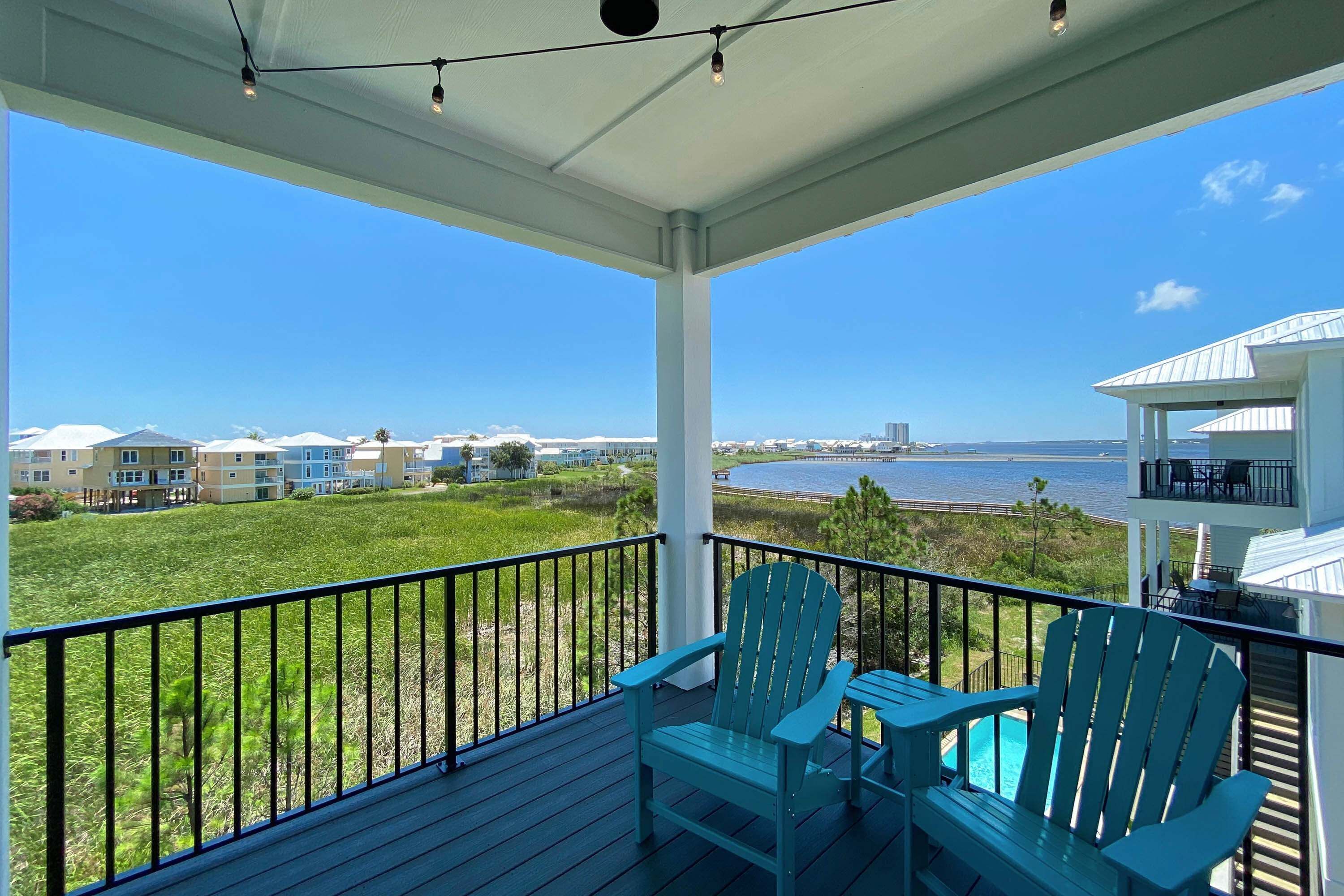 A balcony with two Adirondack chairs and a table, overlooking a grassy area and a body of water with buildings in the background.