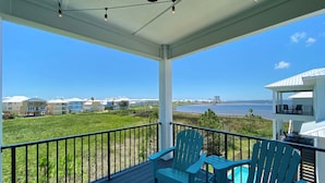 A balcony with two Adirondack chairs and a table, overlooking a grassy area and a body of water with buildings in the background. - Azure Horizon Beachfront Charm in Gulf Shores (Gulf Shores)