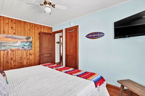 A bedroom with a bed, a ceiling fan, a painting, a wooden door, and a mounted television.