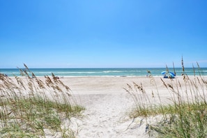 Ein Strand mit blauem Himmel, einem Sandweg zum Wasser und einem blauen Sonnenschirm im Sand.