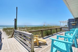 Eine Holzterrasse mit Blick auf den Strand, ausgestattet mit einem Hollerleger, einem gelben Stuhl und einem blauen Stuhl.