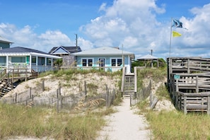 A beachfront property with a sandy path leading to a house, a wooden fence, and a flag.