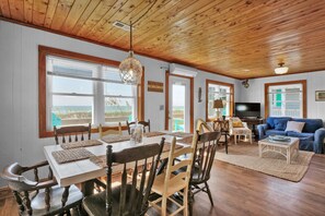 A cozy interior with a wooden ceiling, large windows overlooking a beach, a dining area with a table and chairs, and a blue sofa in the living room. - Seaworthy - Dog Friendly, Updated Classic Cottage with Bonus Space! (Topsail Beach)