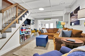 A living room with a staircase, a blue sofa, a brown couch, and a TV on a shelf. - Seaworthy - Dog Friendly, Updated Classic Cottage with Bonus Space! (Topsail Beach)