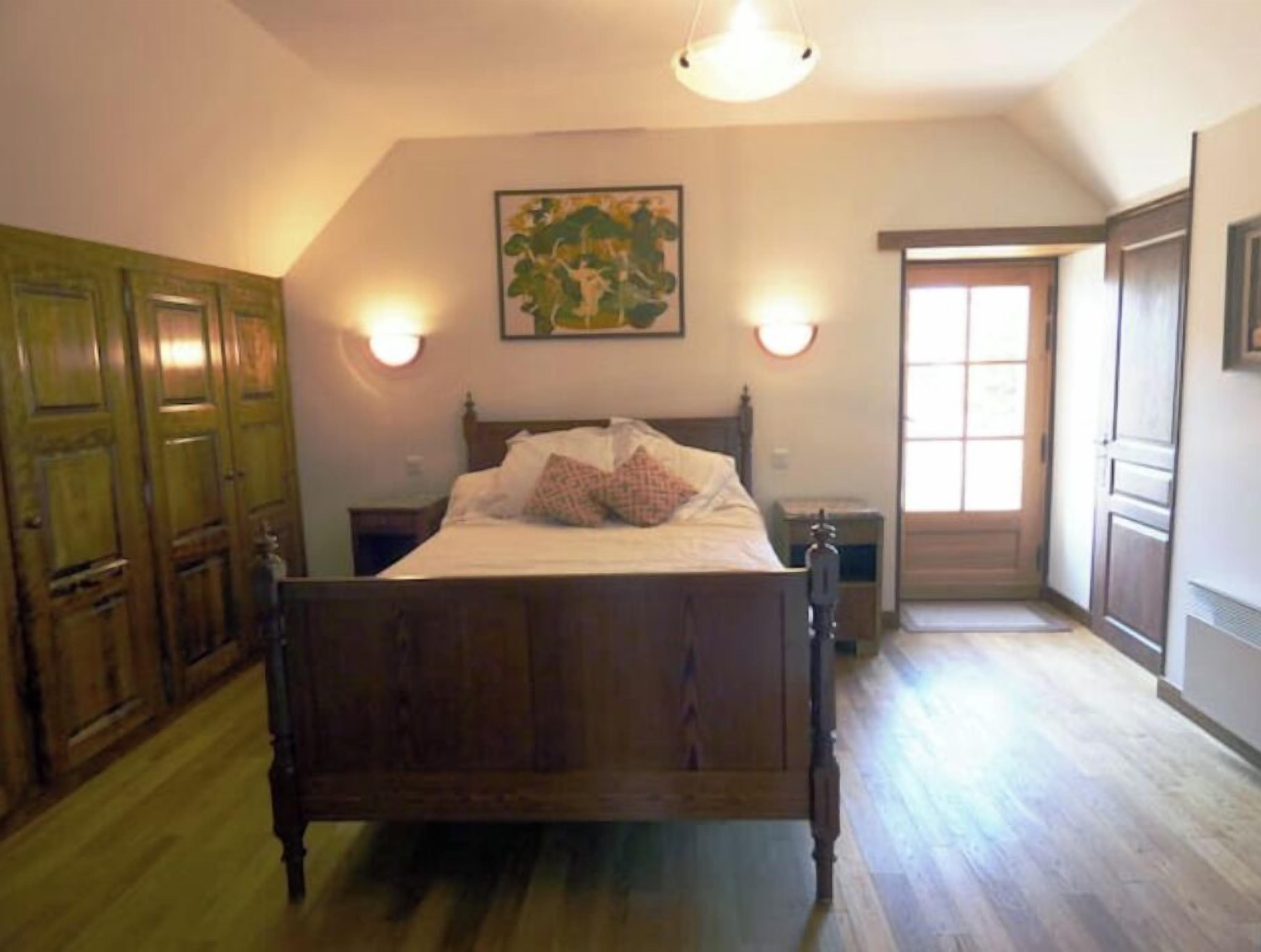 A bedroom with a wooden bed, two bedside tables, a nightstand, a door, and a wall-mounted light fixture.