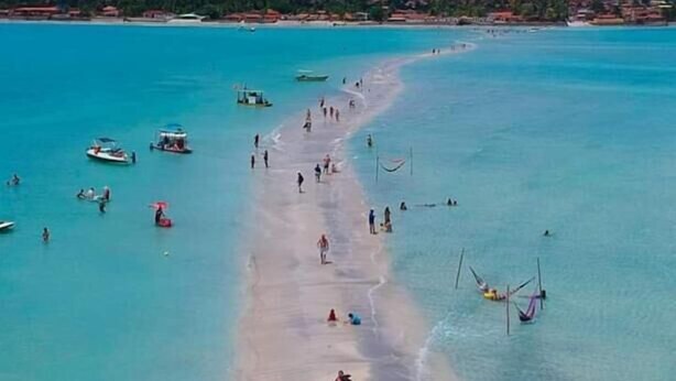 Una playa con agua turquesa cristalina, gente caminando por la orilla y barcos a lo lejos.