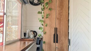 A kitchen with a wooden cabinet, a black chair, a fan, and a window with a view of a red building. - Tuk Tuk Gilhari Noida (Noida)
