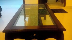 Een tafel met een glazen blad en donker houten poten, in een kamer met een bed en een stoel op de achtergrond.
