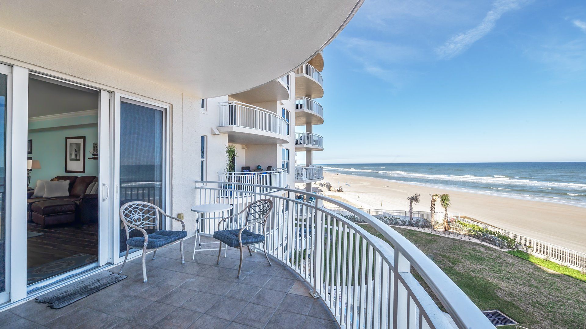 A balcony with a view of the beach, a dining area with chairs, and a living room with a sofa.