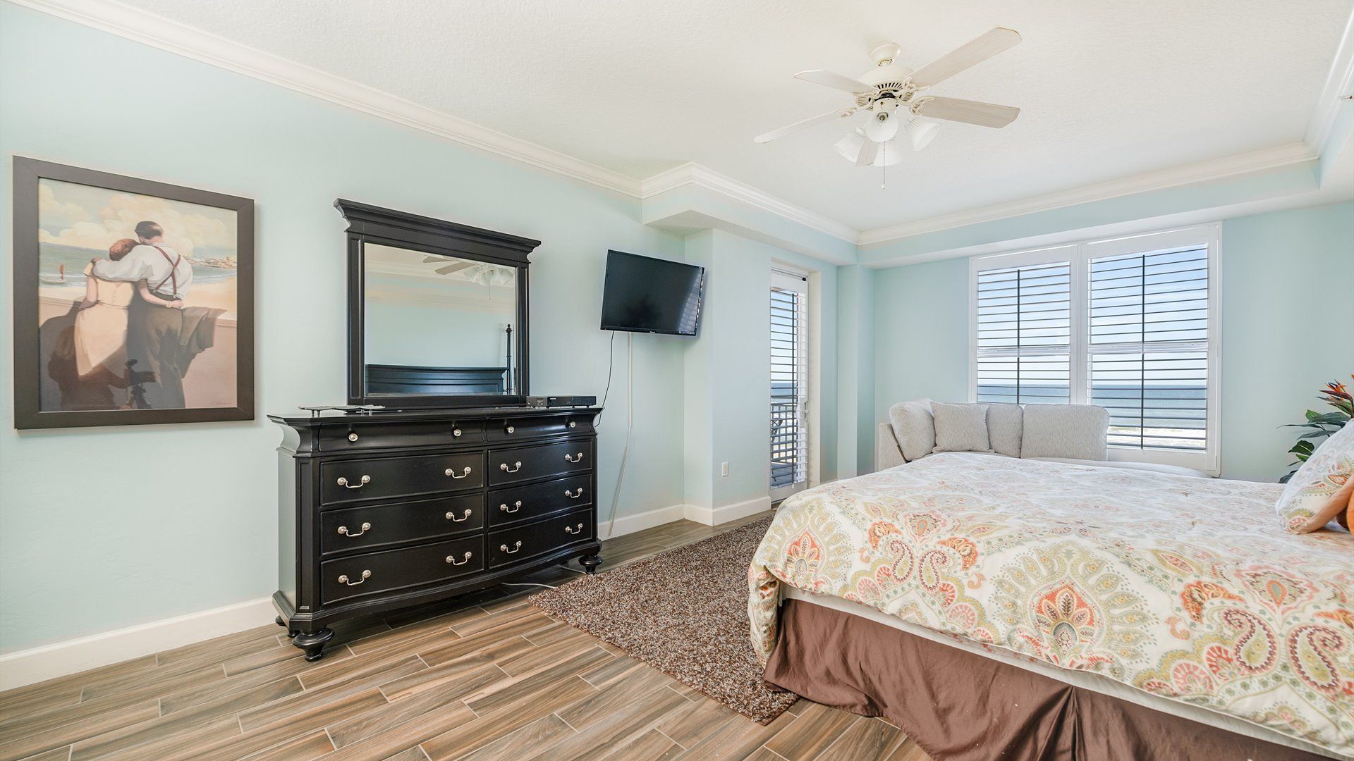A bedroom with a large bed, a dresser, a ceiling fan, a TV, and a painting on the wall.