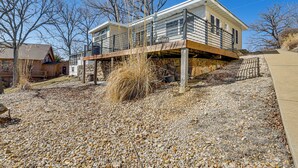 A two-story house with a deck, surrounded by trees and a gravel driveway.