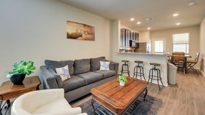 Un salon moderne avec un canapé gris, un fauteuil blanc, une table basse en bois et un coin repas comprenant une table en bois et des tabourets de bar.