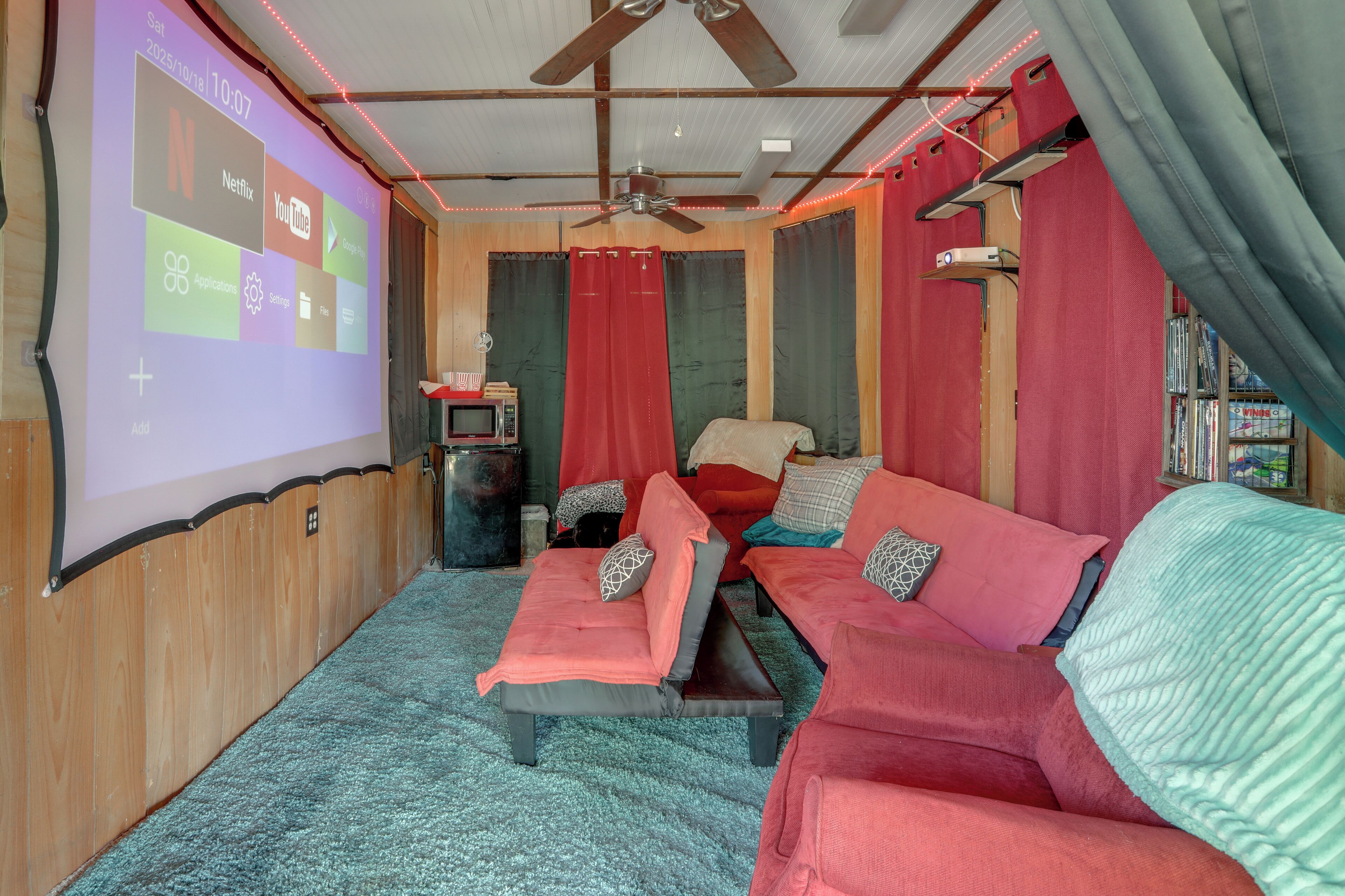 A cozy living room with a large screen displaying streaming services, a ceiling fan, and a bookshelf.