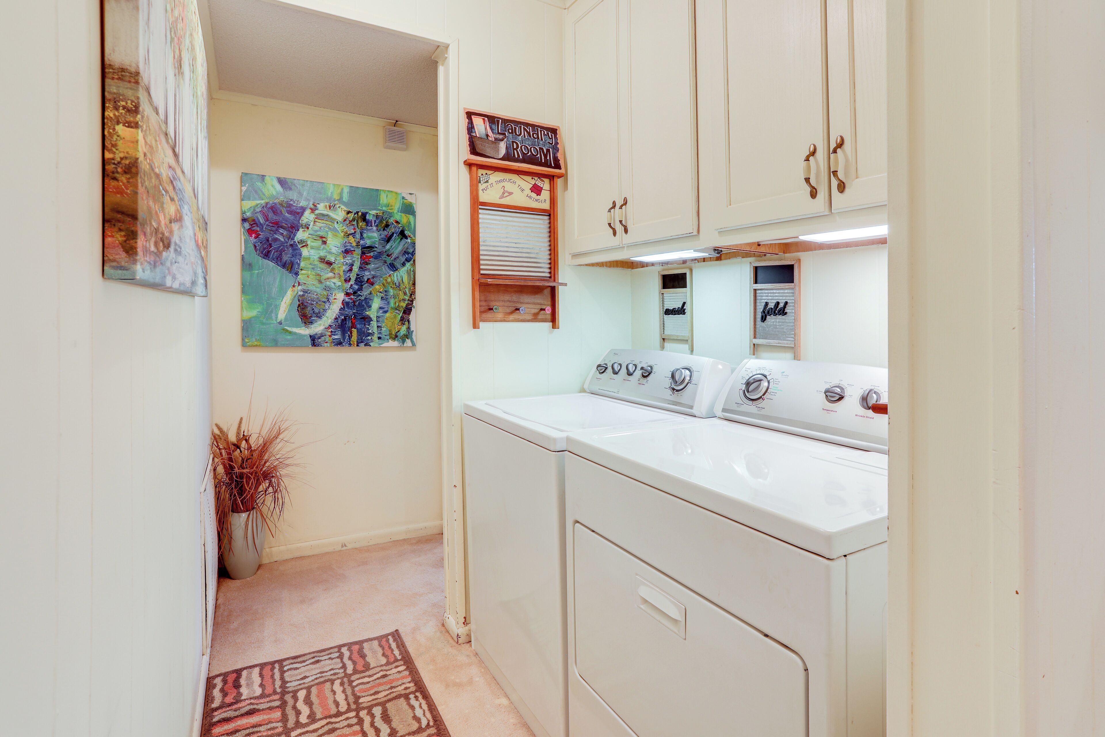 A laundry room with a washing machine, dryer, and a sign indicating it's the laundry room. There is a painting on the wall and a plant in a vase.