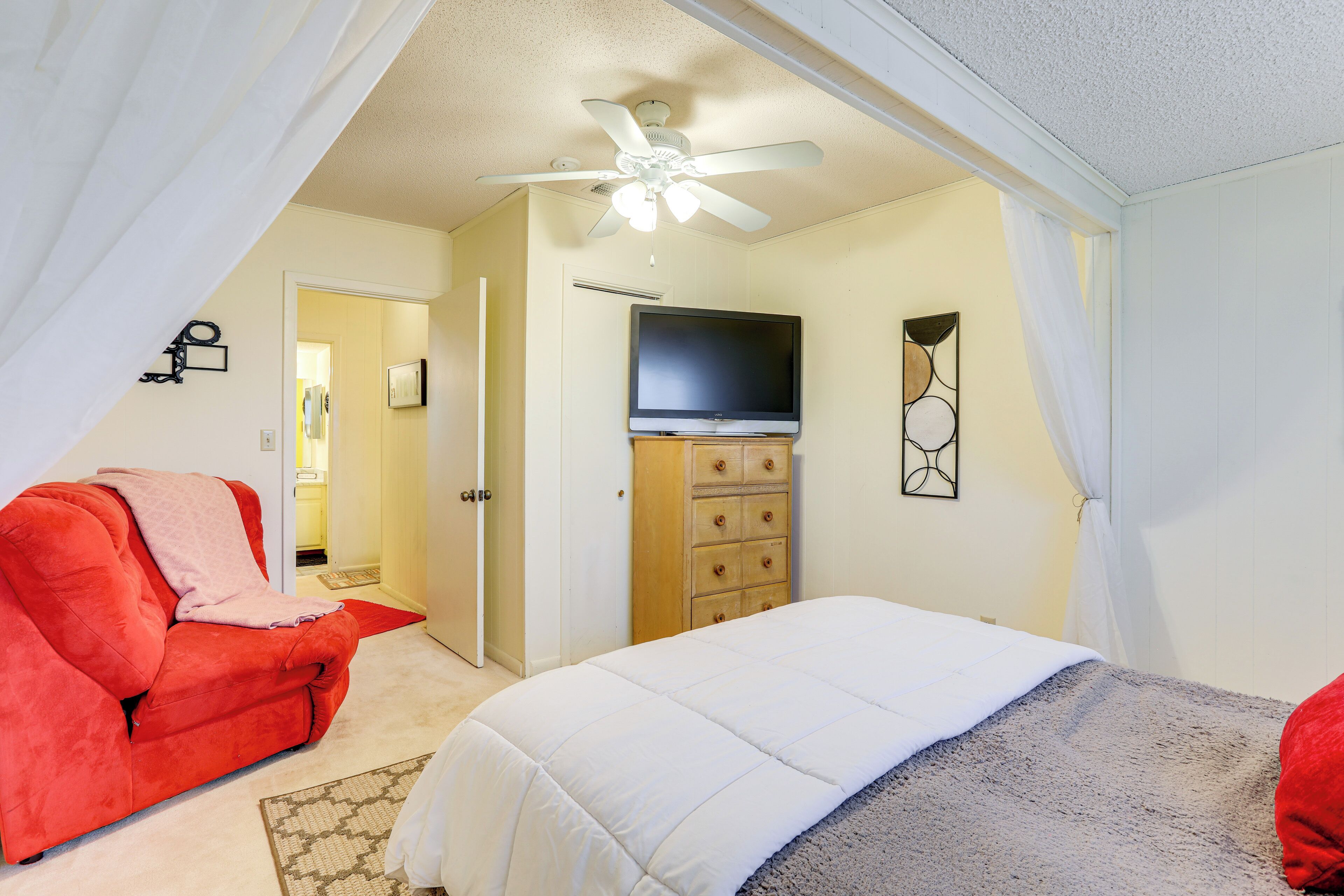 A bedroom with a bed, a red chair, a ceiling fan, a television, and a dresser.