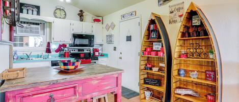 Une cuisine avec des meubles blancs, une table en bois et une chaise rouge. On y trouve des étagères avec des tasses rouges et un panneau indiquant «
Bienvenue
».