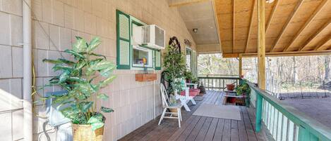 A covered porch with a wooden floor, white walls, and green shutters. There are potted plants, a white chair, and a table with a red cushion.