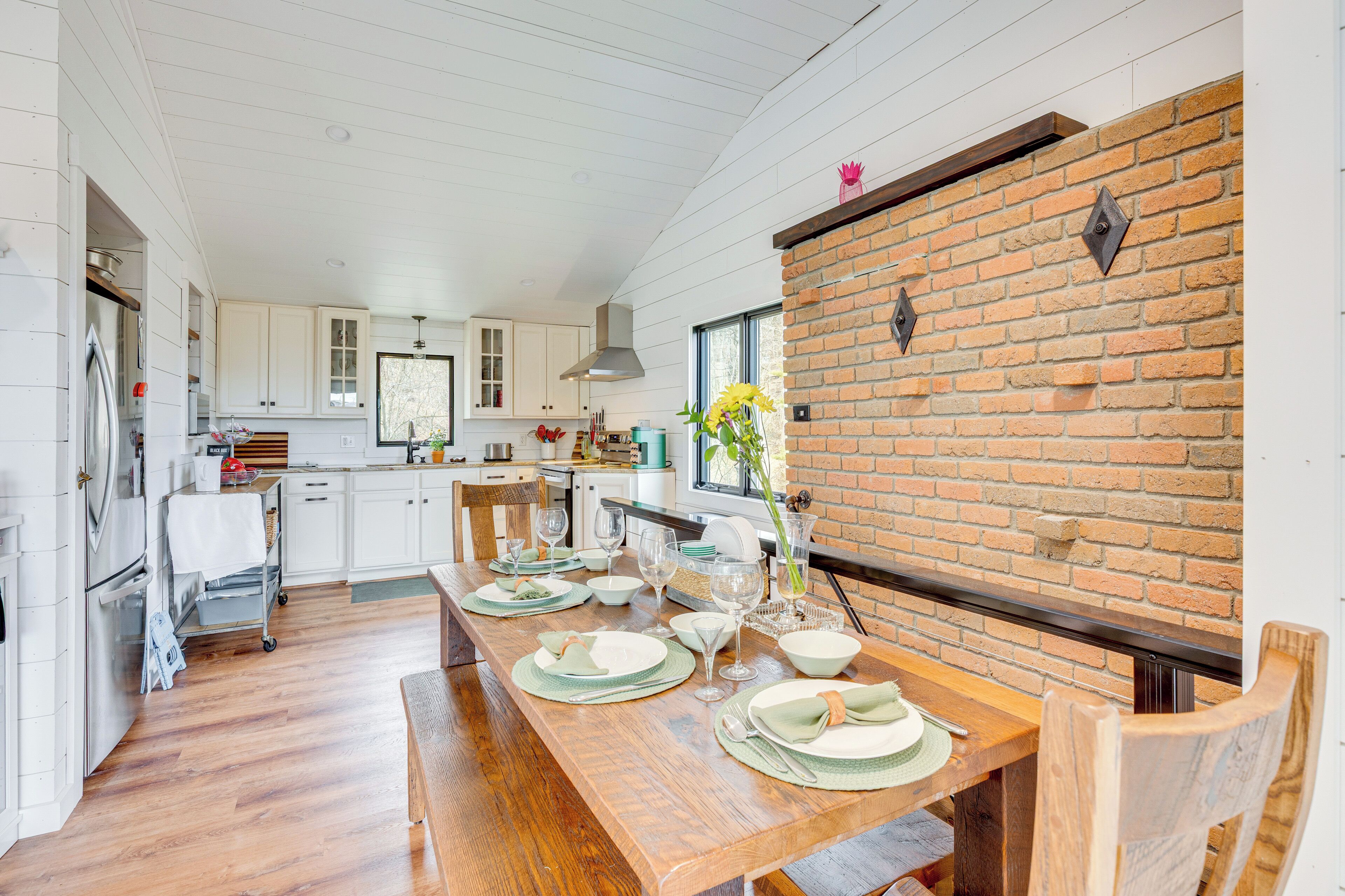 A modern kitchen with white cabinetry, a wooden dining table set for six, and a brick accent wall.