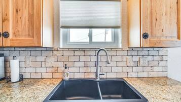 Una cocina con encimera de granito, pileta doble, muebles de madera, una ventana con persiana y un salpicadero de ladrillo.